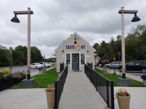 A small building with a sign reading "Taste NY," surrounded by greenery and parked cars under a cloudy sky.