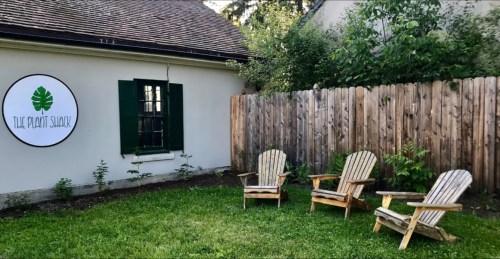 A cozy outdoor space with three wooden chairs on grass beside a wooden fence and a sign for "The Plant Shack."