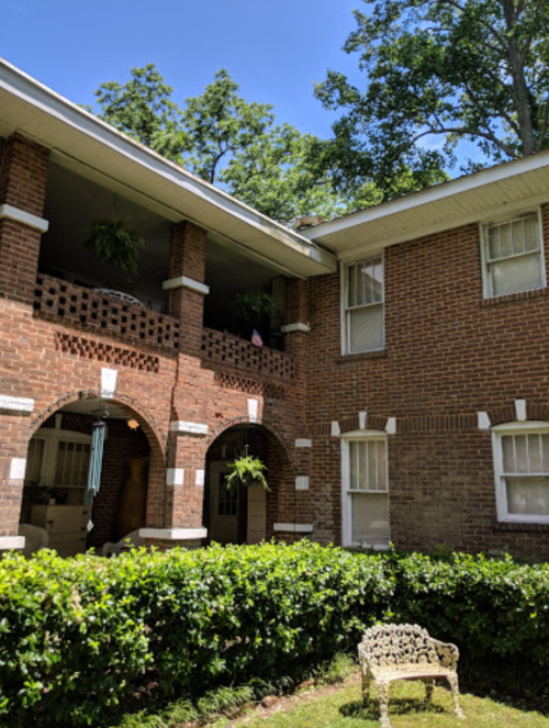 A brick building with a balcony, surrounded by greenery and a small white chair in the foreground.