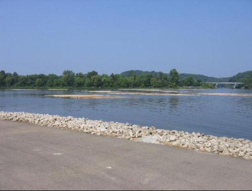 A calm river scene with a rocky shore, green trees in the background, and a clear blue sky.