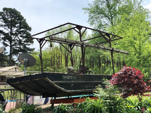 An old boat on a metal frame, surrounded by greenery and a small red tree in a residential area.