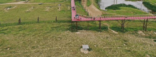 Aerial view of a pink walkway over green fields, with people walking and a small shelter visible below.