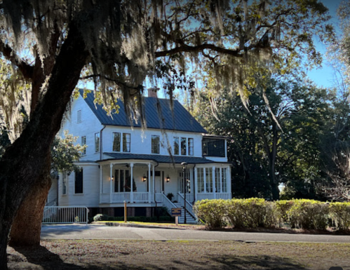 A large white house with a porch, surrounded by trees and Spanish moss, set in a lush green landscape.