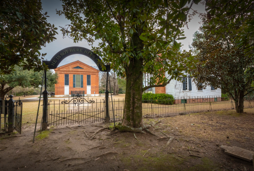 A wrought iron gate with a sign opens to a grassy area featuring two buildings surrounded by trees.