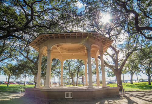 A gazebo surrounded by trees, with sunlight filtering through the leaves and a clear blue sky in the background.