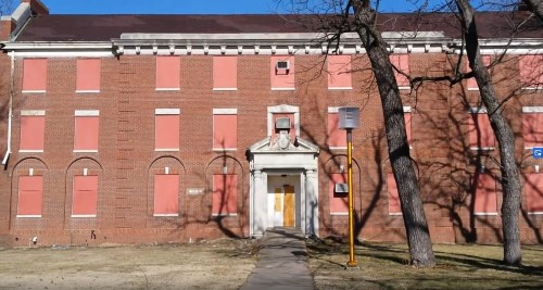 Abandoned brick building with boarded windows, a central entrance, and a tree in front against a clear blue sky.