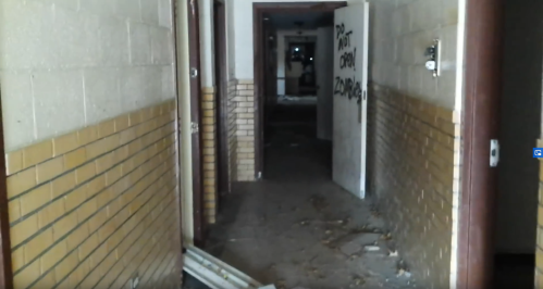 A dimly lit, abandoned hallway with graffiti on the walls and debris on the floor. An open door leads to another room.