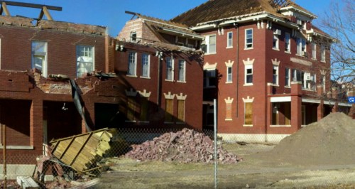 Abandoned brick building with damaged walls and debris, surrounded by a fence and piles of rubble.
