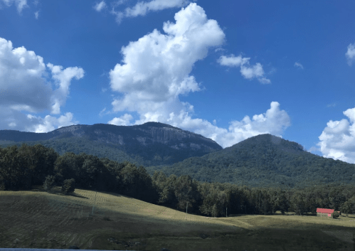 Scenic view of mountains under a blue sky with fluffy clouds, surrounded by green fields and trees.