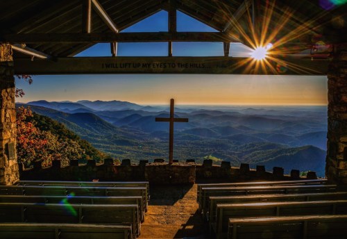 A scenic view from a stone pavilion overlooking mountains, with a cross in the foreground and sunlight shining through.
