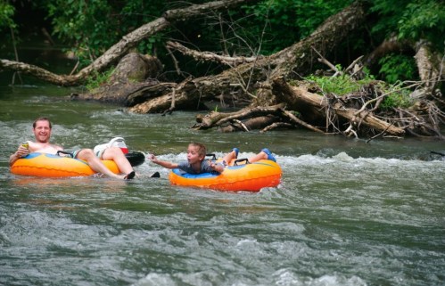 A man and a boy float on orange inner tubes down a river, surrounded by greenery and fallen branches.