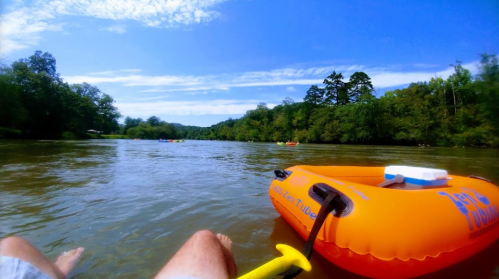 A person relaxing in an orange tube on a calm river, surrounded by trees and blue sky.