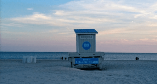 Lifeguard tower at Atlantic Beach with a calm ocean and pastel sky in the background, people walking along the shore.