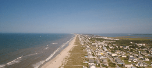 Aerial view of a sandy beach stretching along the coastline, with houses lining the shore and calm blue waters.
