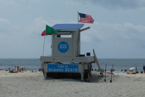 Lifeguard tower at Atlantic Beach with flags, surrounded by beachgoers and umbrellas on a cloudy day.