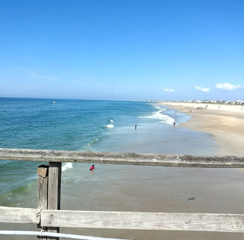 A view of a sandy beach and calm ocean under a clear blue sky, seen from a wooden pier.