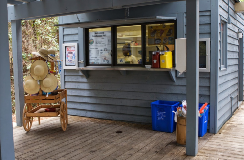 A small wooden kiosk with a menu, serving area, and a cart of straw hats outside on a wooden deck.