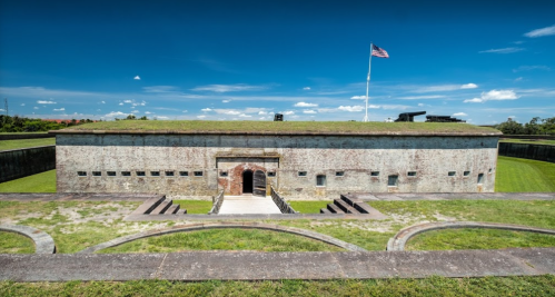 Historic fort with a grassy roof, stone walls, and an American flag flying against a clear blue sky.