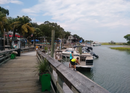 A wooden boardwalk along a calm waterway with boats docked and people enjoying the scenery on a sunny day.