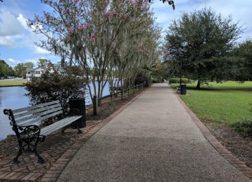 A peaceful park pathway lined with trees, a bench, and a river, under a partly cloudy sky.