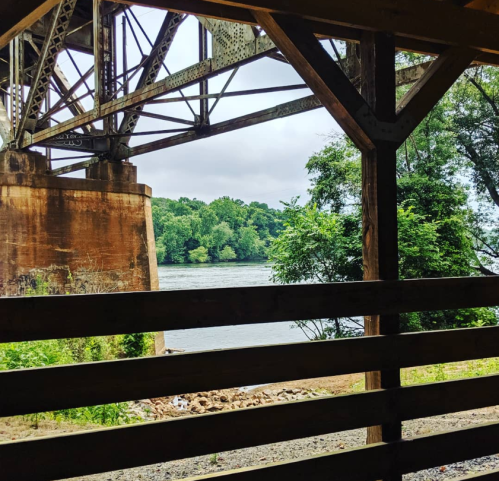 View of a river framed by wooden beams and a bridge, surrounded by lush greenery under a cloudy sky.