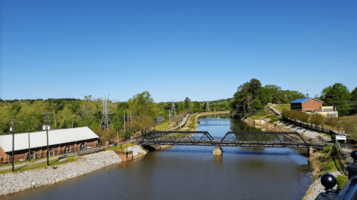 A serene river scene with a bridge, green trees, and a clear blue sky, alongside a walking path and buildings.