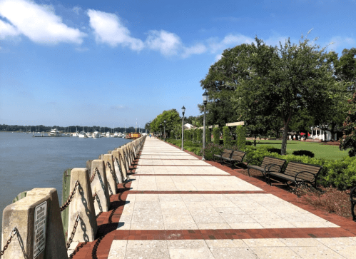 A scenic waterfront path lined with benches and trees, overlooking a calm river with boats in the distance.