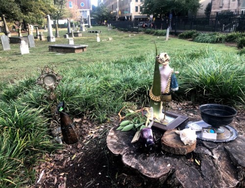 A small altar with various items, including a statue, bottle, and flowers, set in a grassy area near gravestones.