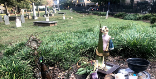 A cemetery scene with gravestones, overgrown grass, and a decorative statue among plants and offerings.