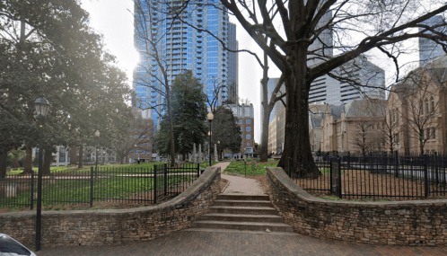 A park with trees and a stone pathway, surrounded by tall buildings and a fence, under a clear sky.