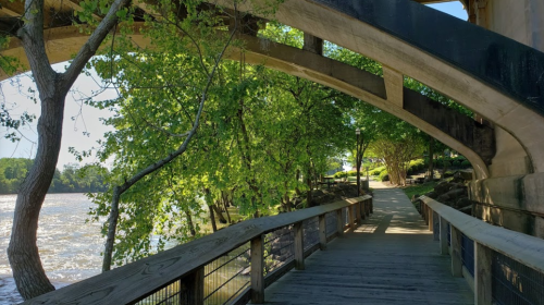 A wooden walkway under a curved bridge, surrounded by lush green trees and a river in the background.