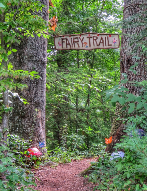 A whimsical entrance to a forest path marked "Fairy Trail," surrounded by lush greenery and colorful decorations.