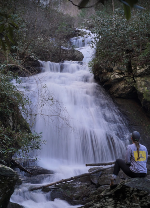 A person sits on rocks by a cascading waterfall, surrounded by lush greenery in a serene natural setting.