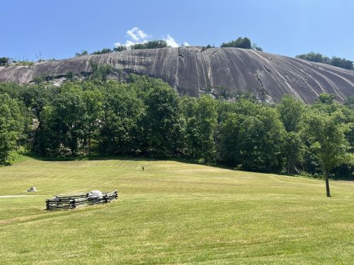 A grassy field with a wooden fence in the foreground and a large, smooth rock face rising in the background under a blue sky.