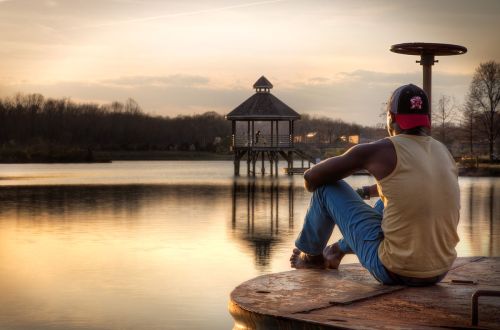 A person sits on a dock by a calm lake at sunset, gazing at a gazebo in the distance.