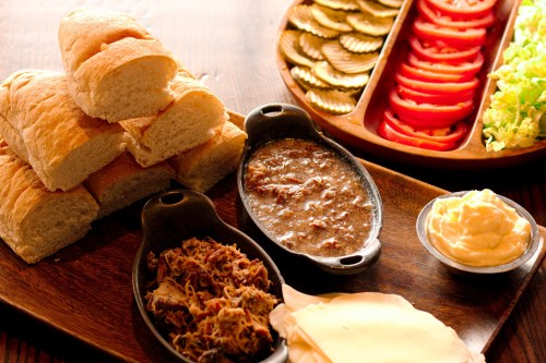 A wooden platter with sliced bread, pulled meat, sauces, and fresh vegetables like pickles, tomatoes, and lettuce.