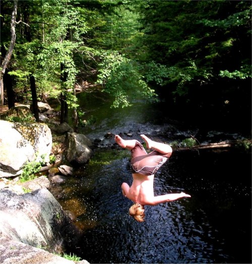 A person performs a backflip into a serene, wooded swimming hole surrounded by rocks and trees.
