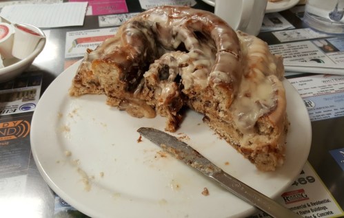 A partially eaten cinnamon roll on a plate, with icing and a knife beside it, set on a colorful table.
