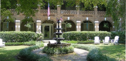 A charming courtyard with a fountain, surrounded by greenery and white chairs, featuring brick arches and American flags.