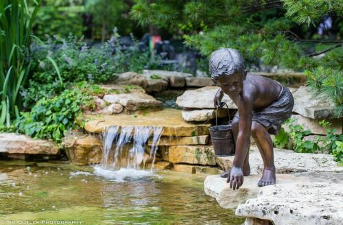 A bronze statue of a boy crouching by a stream, holding a bucket, surrounded by greenery and a small waterfall.