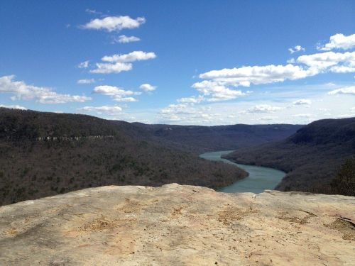 A scenic view of a river winding through a valley, surrounded by hills under a blue sky with fluffy clouds.