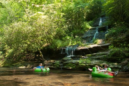 Two people relax on inflatable tubes in a serene stream, surrounded by lush greenery and a small waterfall.