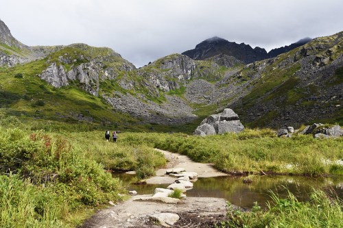 Two hikers walk along a rocky path through a lush green valley surrounded by mountains under a cloudy sky.