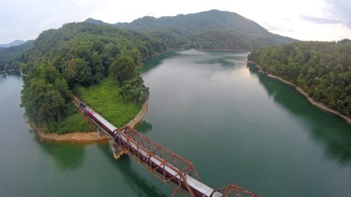 Aerial view of a bridge over a calm lake, surrounded by lush green hills and trees under a cloudy sky.