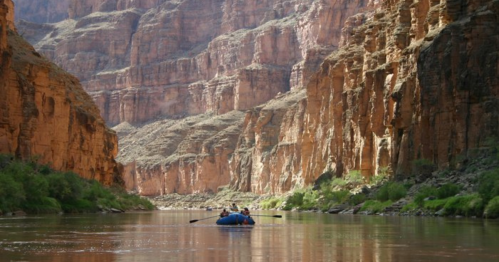 A group of people rafting on a river surrounded by towering red rock cliffs and lush greenery.