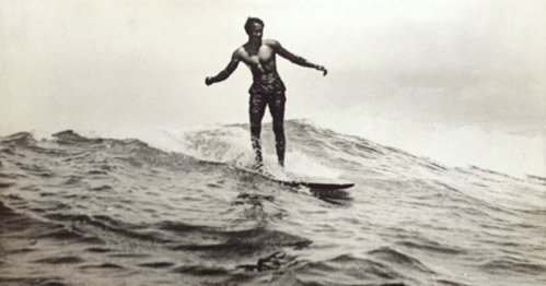 A black and white photo of a man surfing on a wave, balancing on a surfboard with a confident stance.