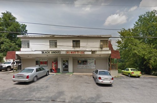 A small deli named "Black Angus" with a sign for deli meats, featuring parked cars in front and trees in the background.