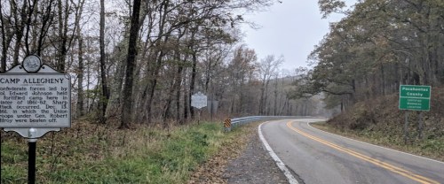 A winding road through a wooded area with historical signs for Camp Allegheny and Pocahontas County.