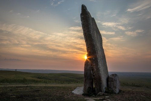 A tall stone monolith stands against a sunset, casting a shadow over a grassy landscape.