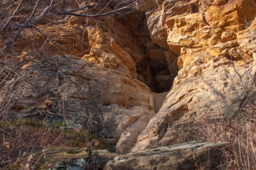 A rocky cliff with layered orange and beige stone, featuring a narrow crevice and sparse vegetation in the foreground.